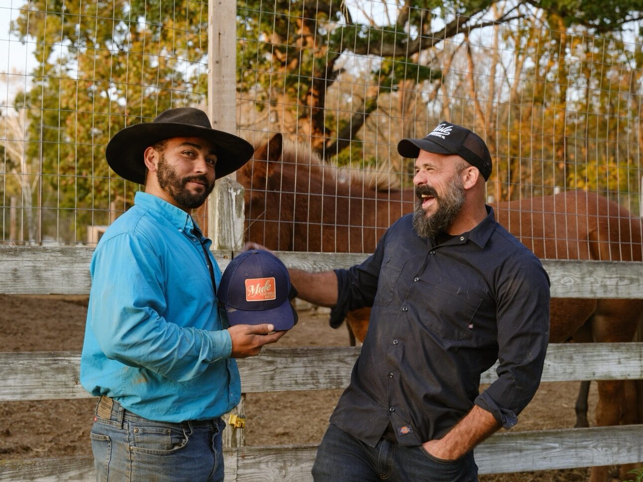 Two men stand by a wooden fence with a horse behind them. One in a cowboy hat and blue shirt holds out a cap, while the other in a baseball cap and dark shirt smiles widely. Behind them, Parr Mule grazes under the trees—true fans of Mule Resonator Guitars.