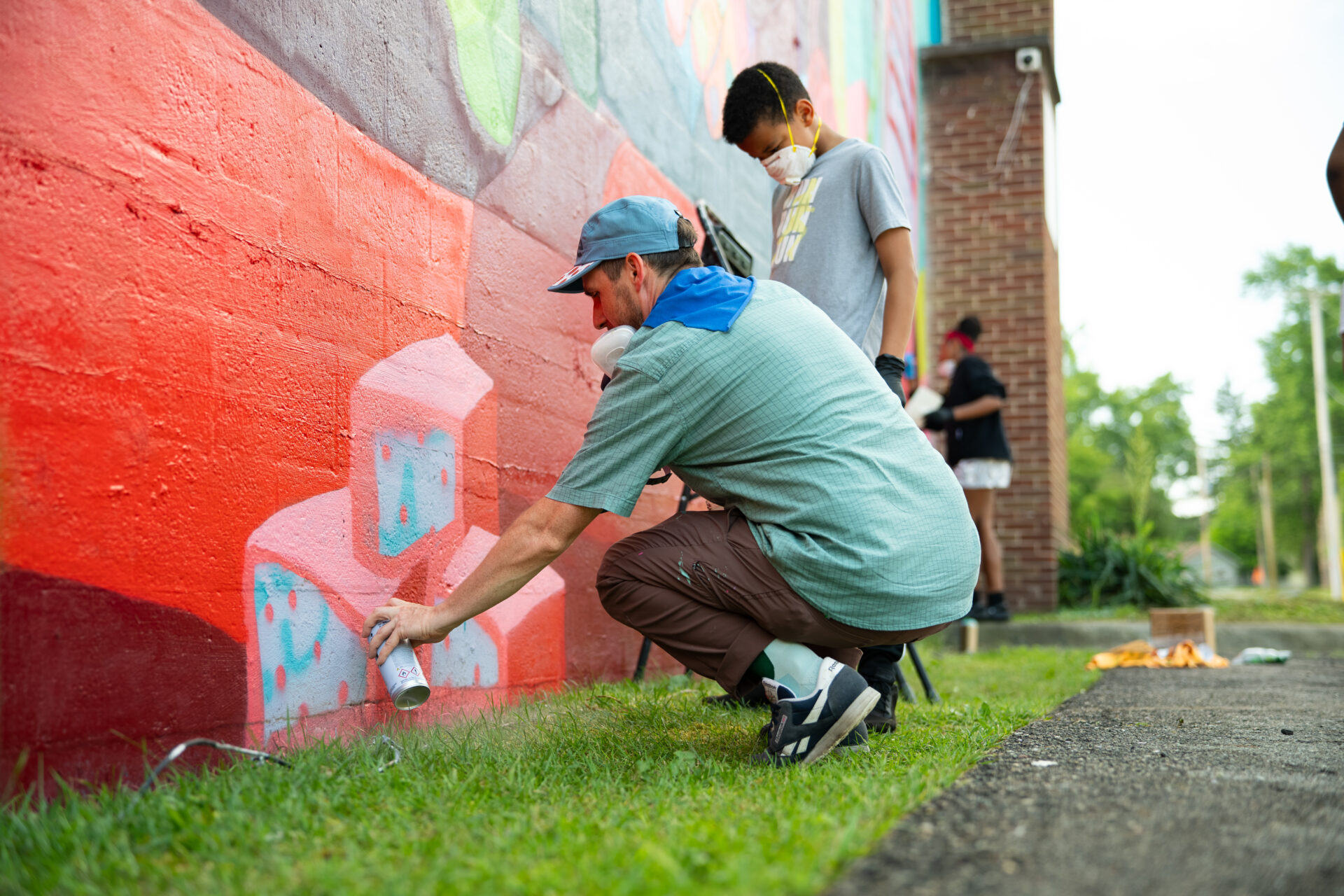 A person crouches and spray-paints colorful shapes on a red wall while another person stands behind, both wearing masks. They are outdoors near grass, with more people and painting supplies in the background.