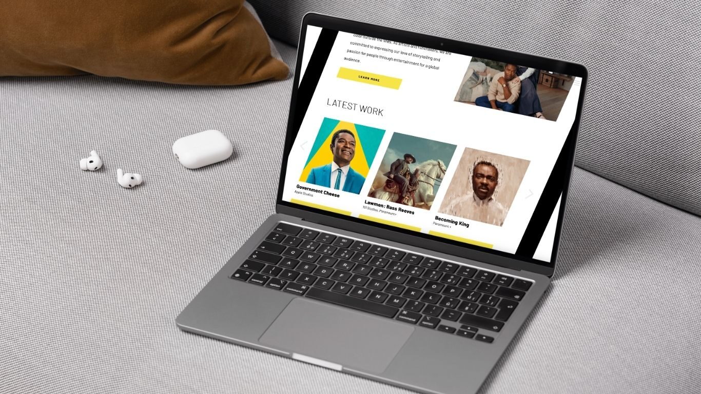 A laptop on a grey couch displays a website featuring highlighted Saxon projects with photos and brief descriptions. Nearby are wireless earbuds and their case on the couch.