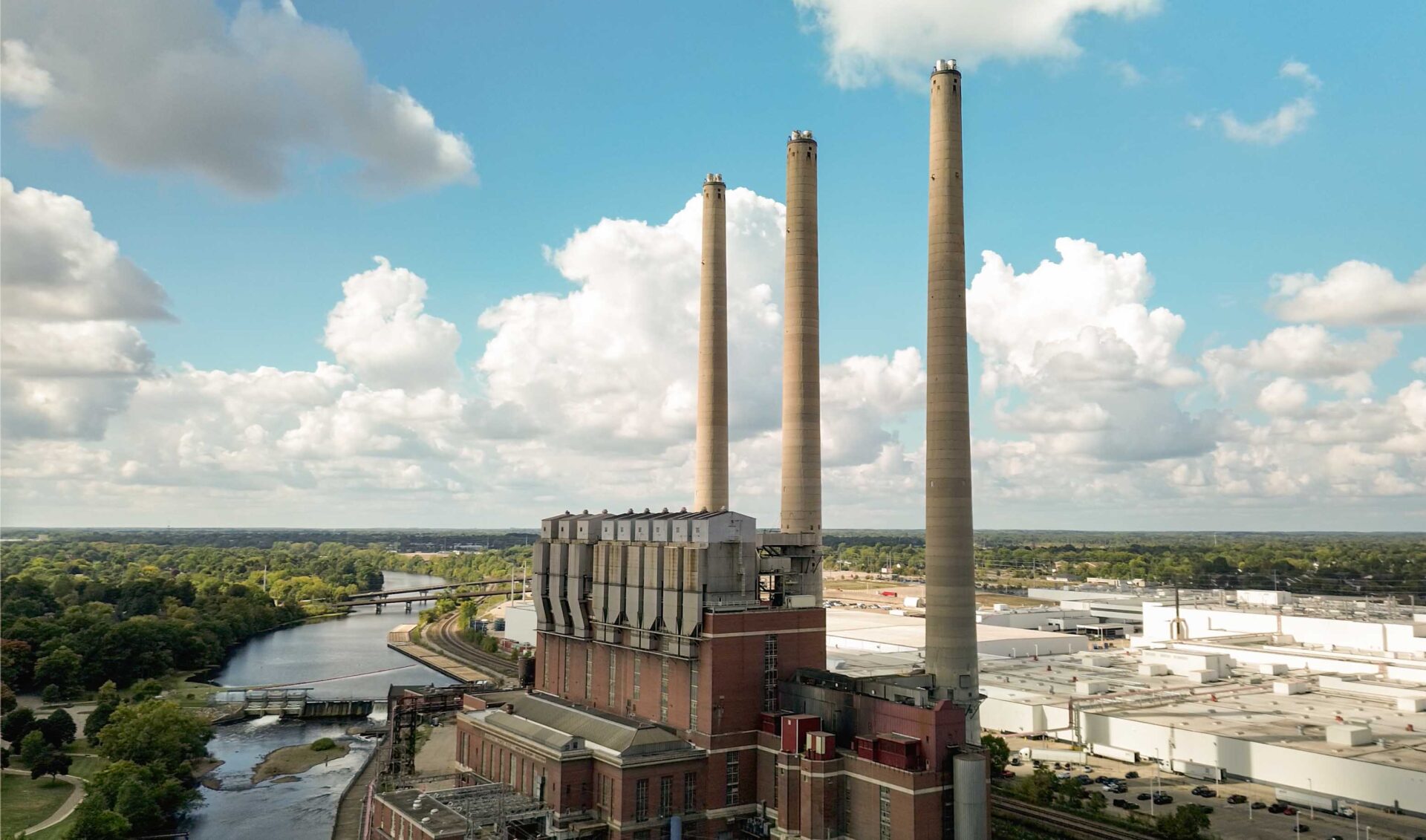 An industrial building with three tall smokestacks stands near a river, surrounded by trees, roads, and warehouses under a partly cloudy sky.