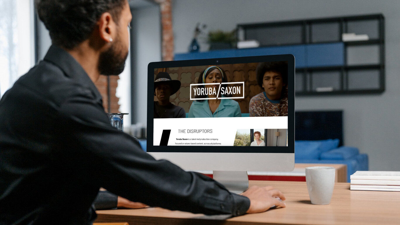 A person sits at a desk using a desktop computer displaying a website titled Yoruba/Saxon with images of three people and a section labeled The Disruptors. A mug and notepad are on the desk.