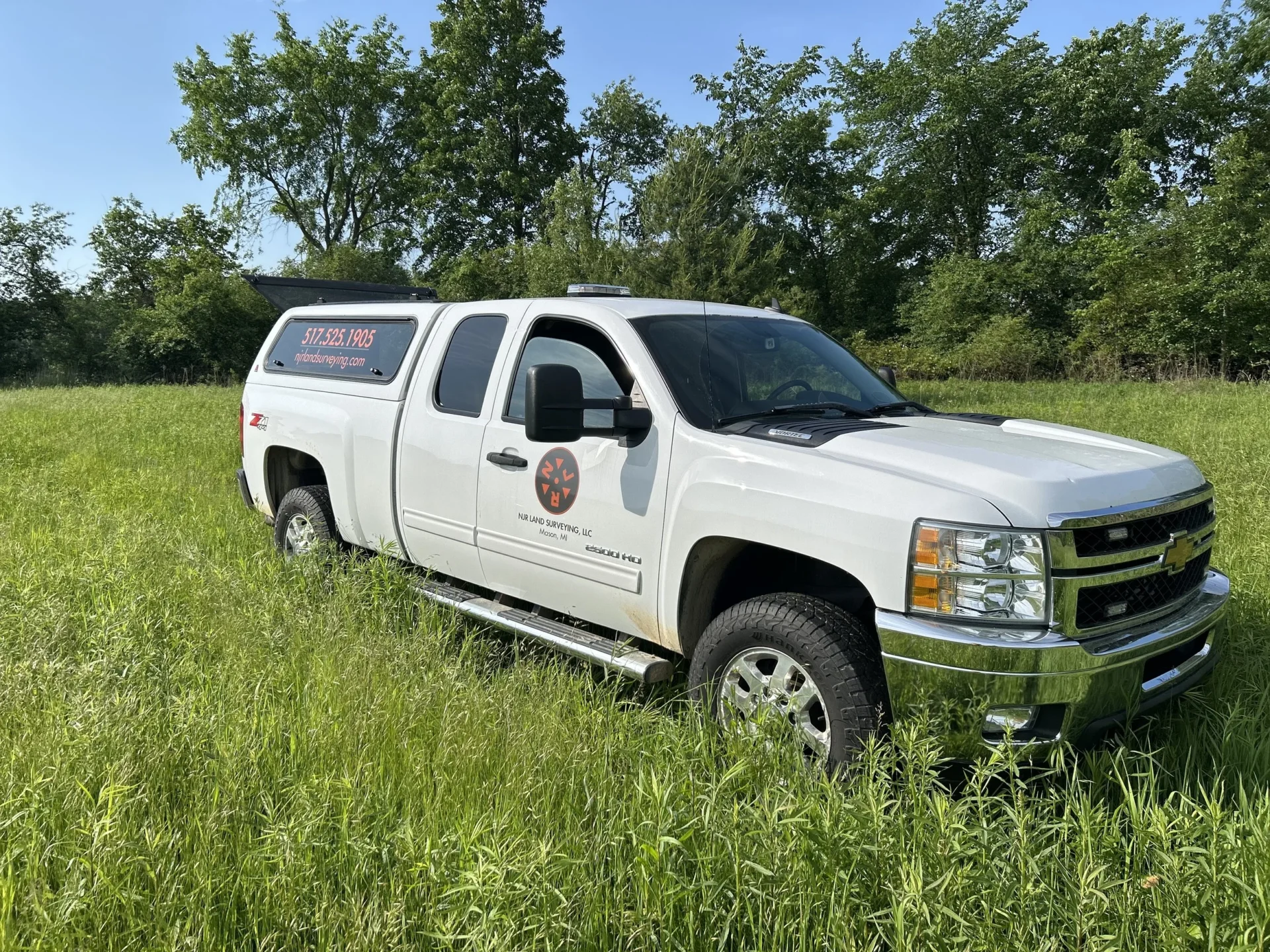 A white NJR Triage pickup truck with a camper shell and company logo is parked in a grassy field, surrounded by green trees under a clear blue sky. The truck displays a phone number, website, and highlights its SEO services on the side.