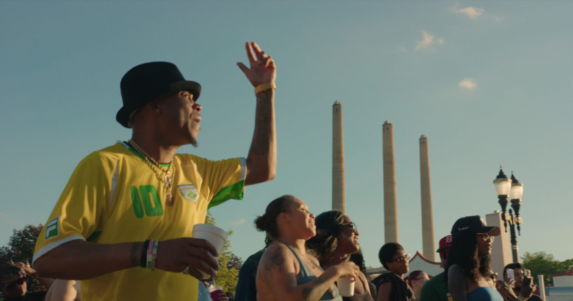 A group of people stands outdoors in sunlight at a Juneteenth Celebration, some smiling and raising their arms. A man in a yellow jersey and black hat holds a cup. Three tall columns and a lamppost stand against the blue sky.