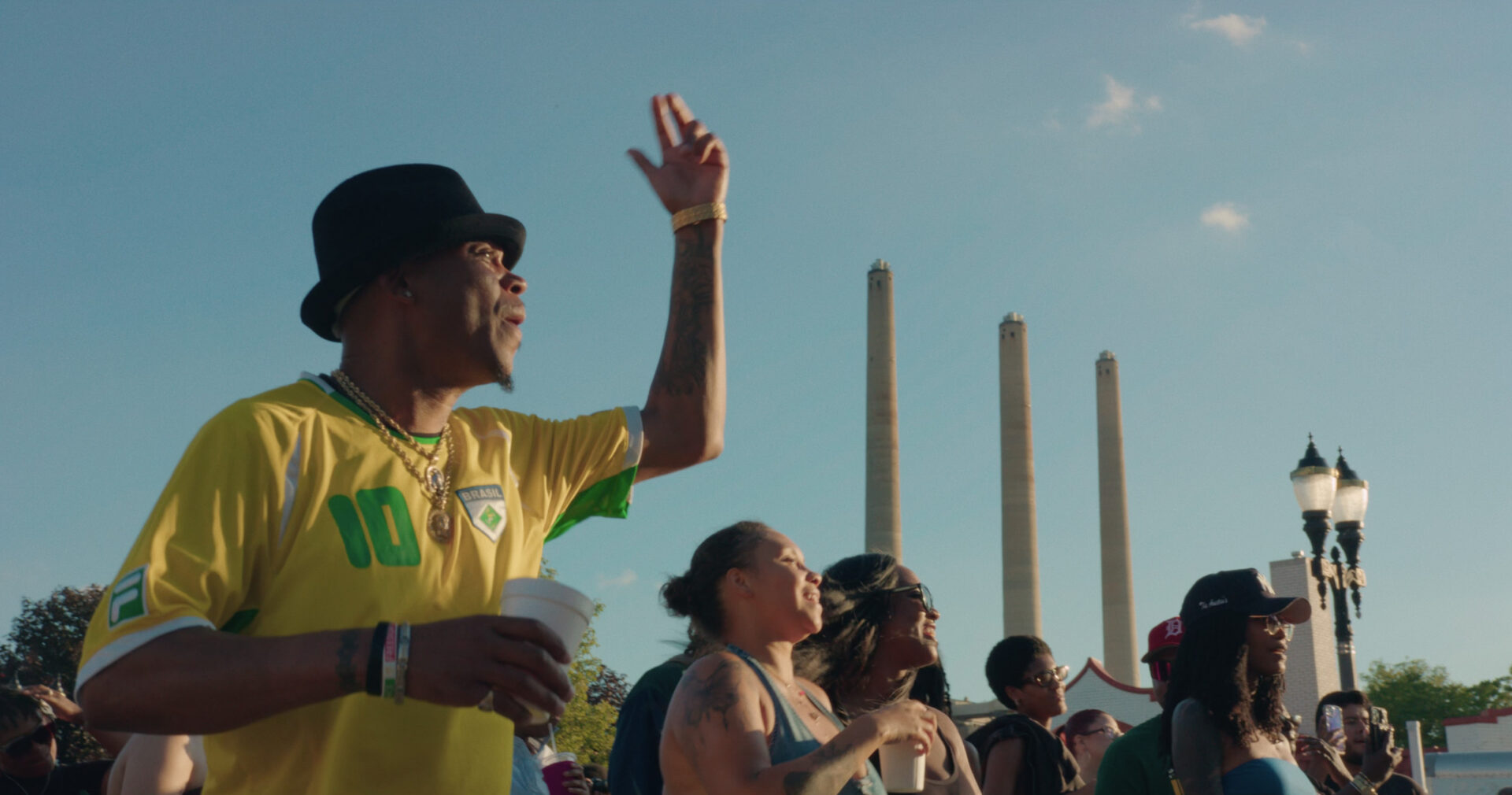 A group of people outdoors at a Juneteenth Celebration watch something off-camera; one man in a yellow Brazil soccer jersey and black hat raises his arm with a cup, while others look on under a blue sky with tall towers in the background.