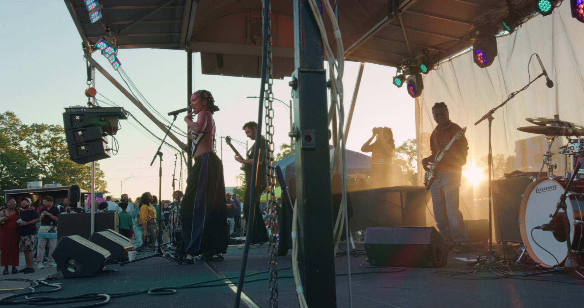 A band performs on an outdoor stage at sunset during a Juneteenth Celebration, with a singer at the front, musicians playing guitar and drums, colorful stage lights, and a crowd gathered in the background.
