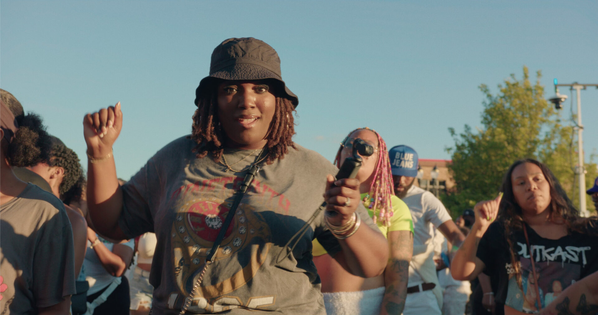 A person in a bucket hat and graphic t-shirt dances joyfully at a vibrant Juneteenth Celebration, holding a microphone among a lively crowd under a clear blue sky.