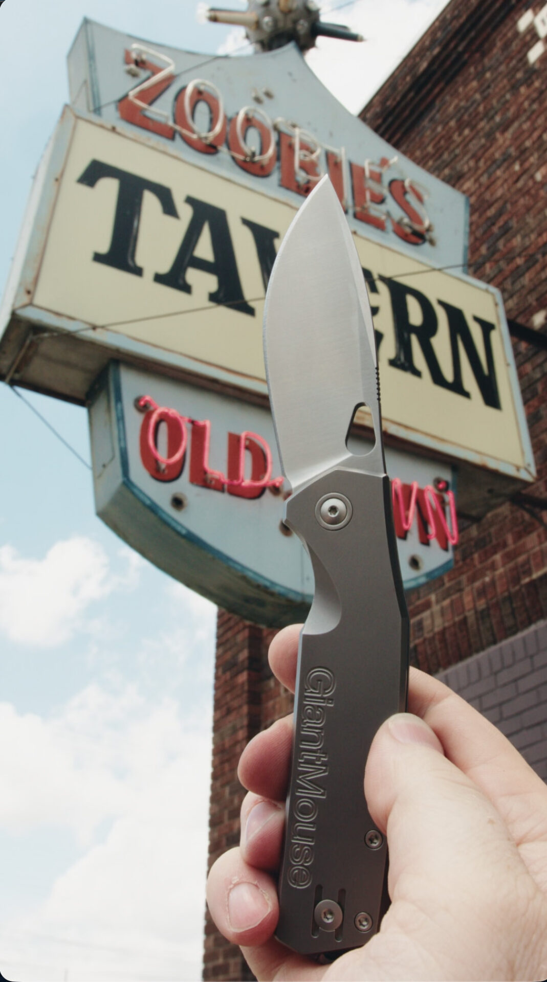 A hand holds an open GiantMouse Knives folding knife in front of a retro sign that reads Zoolies Tavern Old Town attached to a brick building, with a partly cloudy sky—a perfect Zoobie scene for your next social video.
