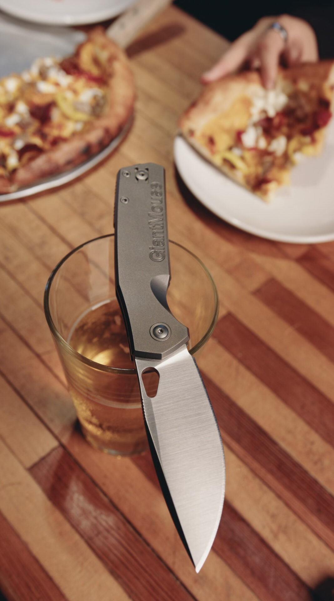 A GiantMouse Knives folding knife balanced on a glass of amber liquid sits on a striped wooden table. In the background, someone holds a pizza slice near a Zoobie, with a pizza tray and more slices visible—perfect for your next social video.
