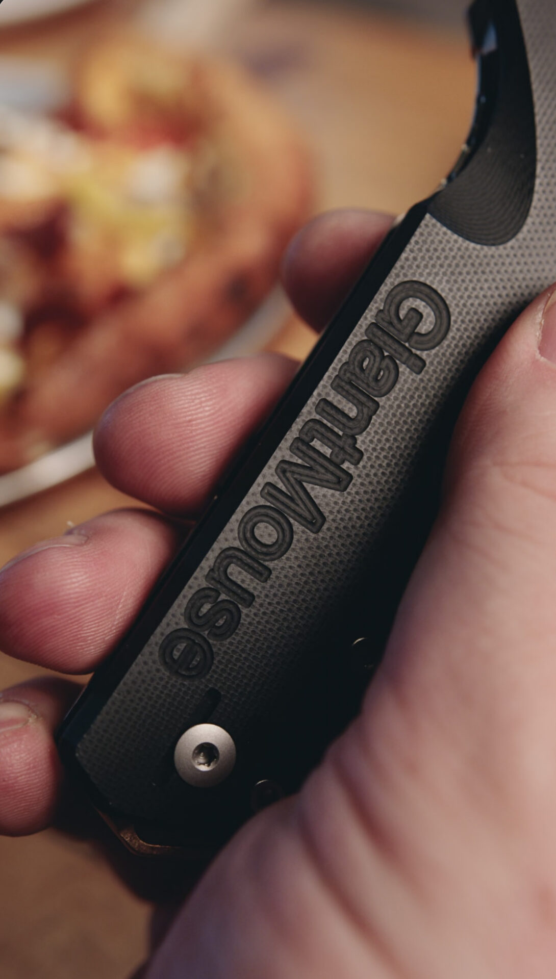 A close-up of a hand holding a black handle with the embossed word Granhaus, reminiscent of premium GiantMouse Knives. The blurred background shows what appears to be food on a plate.