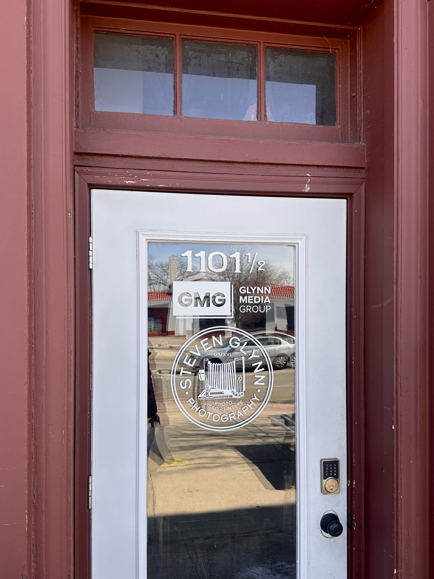 A glass door with the address 1101 1/2 and logos for Glynn Media Group and Steven Glynn Photography, set in a red frame. The door has a keypad lock and reflects parked cars and buildings across the street.