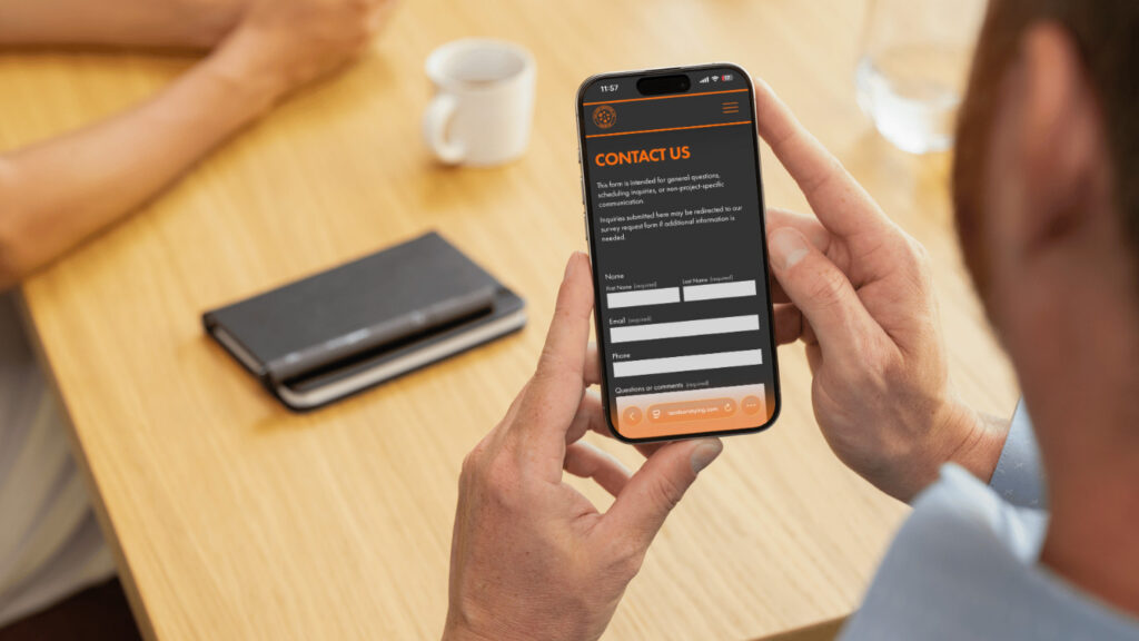 A person holding a smartphone displays a Contact Us form on the screen at a wooden table, with another person’s arm, a notebook, and a glass of water visible in the background.