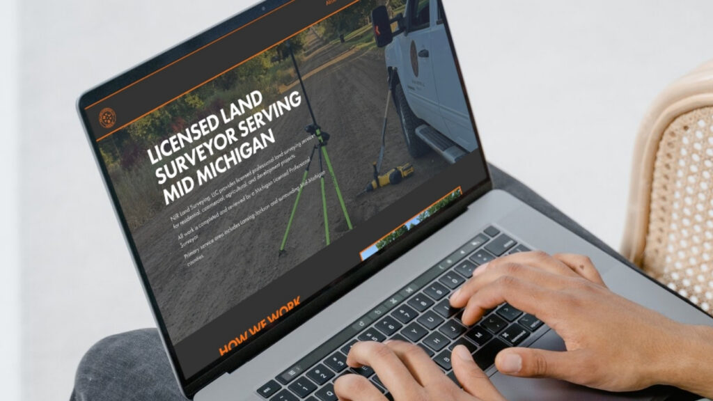 A person types on a laptop displaying a website for a licensed land surveyor serving mid Michigan, with surveying equipment and a white vehicle shown on the site.