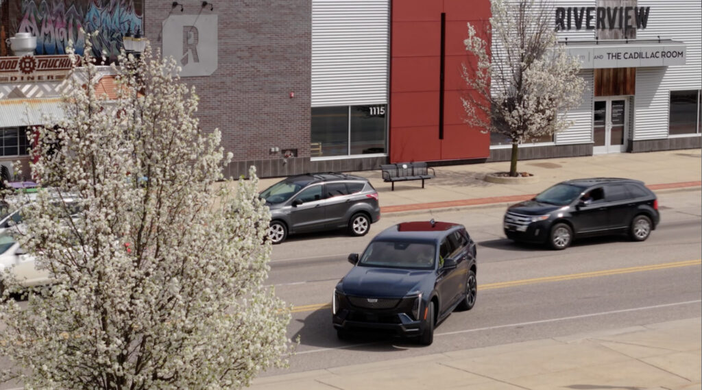 Three cars, including a classic Cadillac, drive down a street lined with blooming trees. A red and gray building and a “RIVERVIEW” sign appear in the background, while a bench sits on the sidewalk—another scene in my Cadillac story.