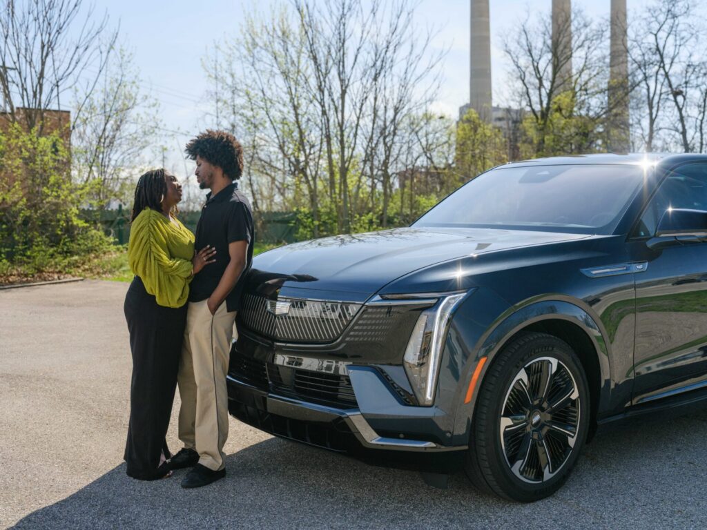 A couple stands close together, smiling at each other near the front of a dark-colored Cadillac in a sunny outdoor setting with trees and industrial chimneys in the background—capturing their unique Cadillac story.