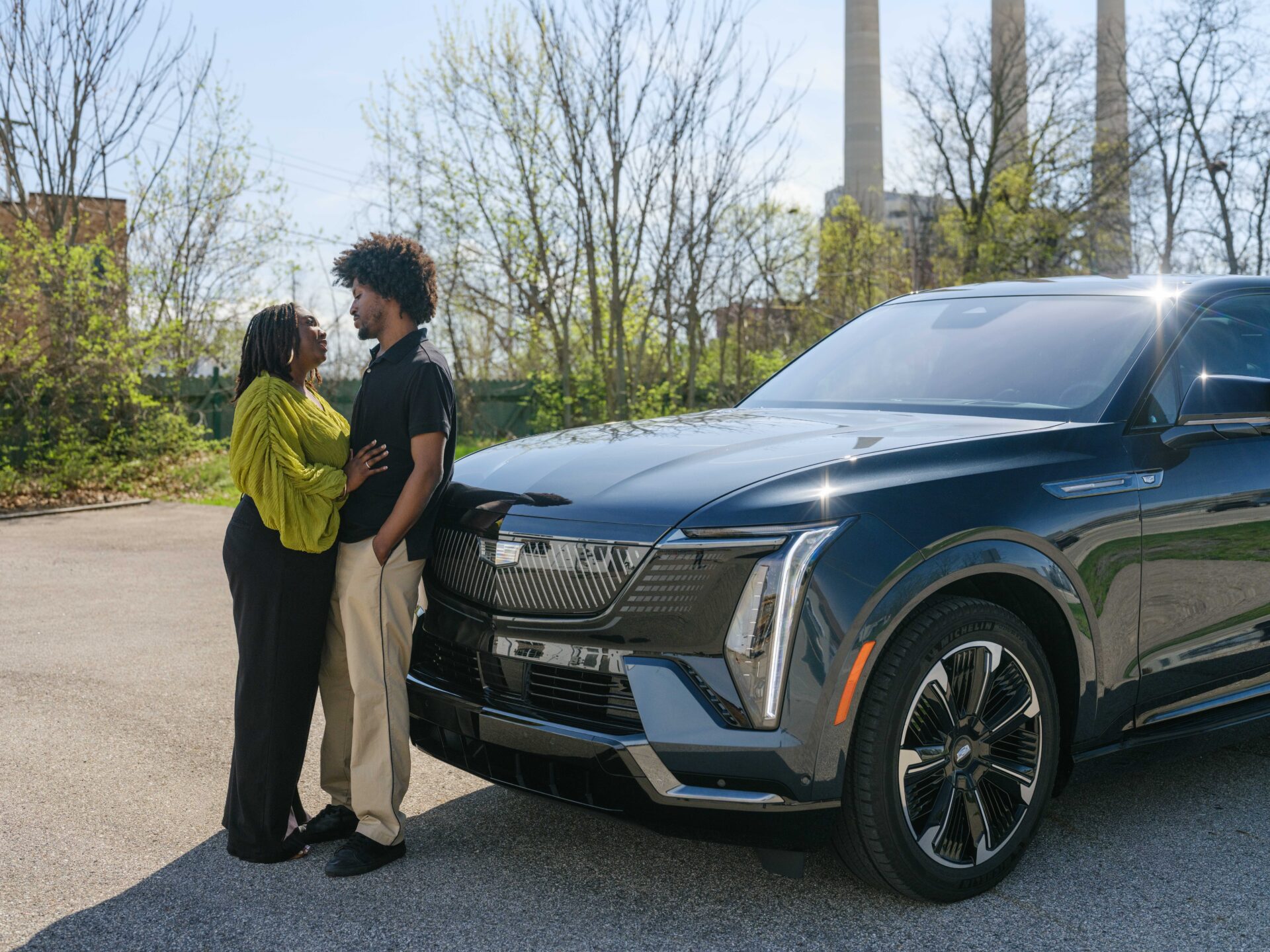 A couple stands close together, smiling at each other near the front of a dark-colored Cadillac in a sunny outdoor setting with trees and industrial chimneys in the background—capturing their unique Cadillac story.