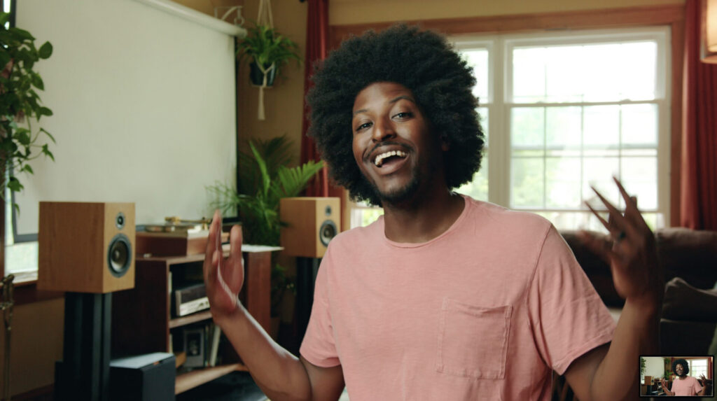 A smiling person with an afro hairstyle wearing a pink t-shirt gestures with both hands while sitting in a cozy, well-lit living room. Creative Sound Solutions speakers and a video call screen are visible in the corner.