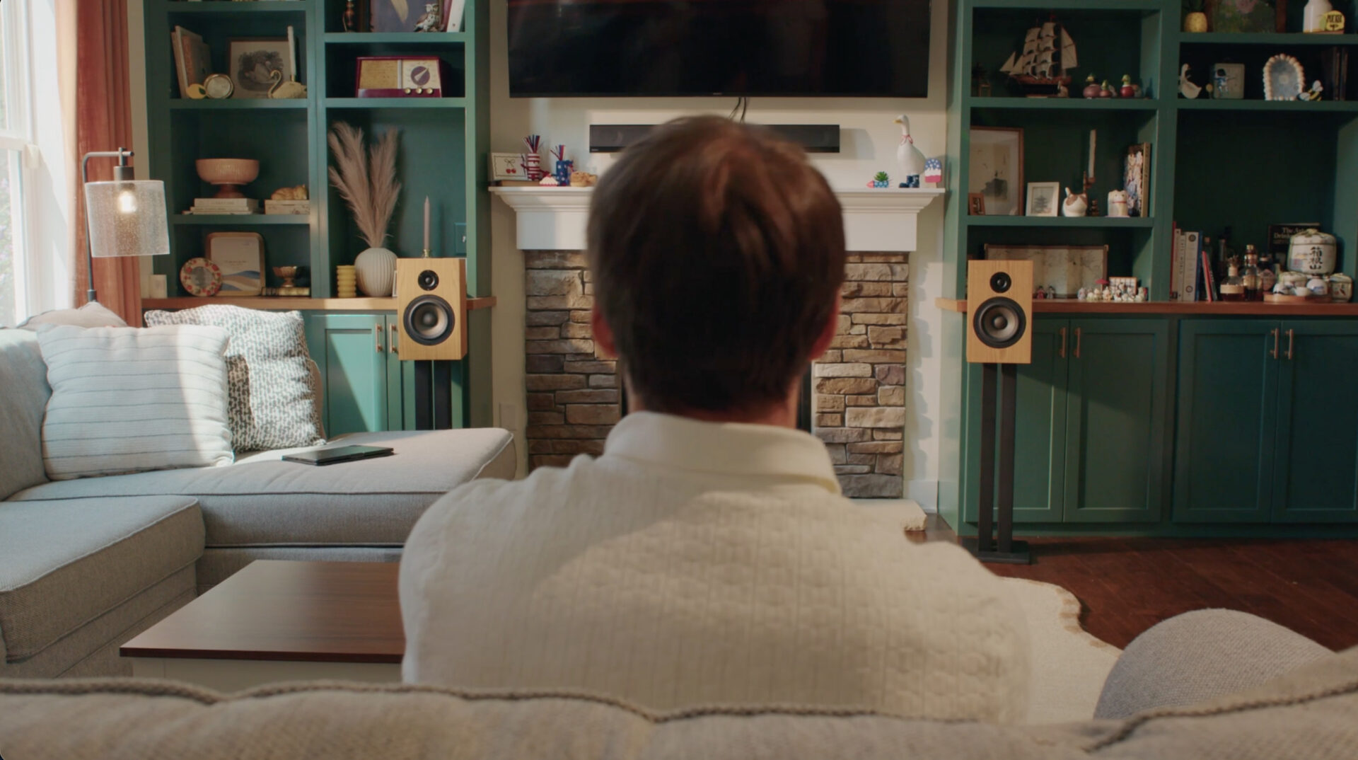 A person sits on a sofa facing a living room with green shelves, a fireplace, speakers, and a TV mounted above the mantel. Decorations and books are arranged on the shelves.